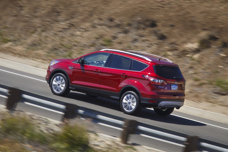Picture of a driving 2017 Ford Escape Titanium in Ruby Red Metallic Tinted Clearcoat from a rear left three-quarter perspective